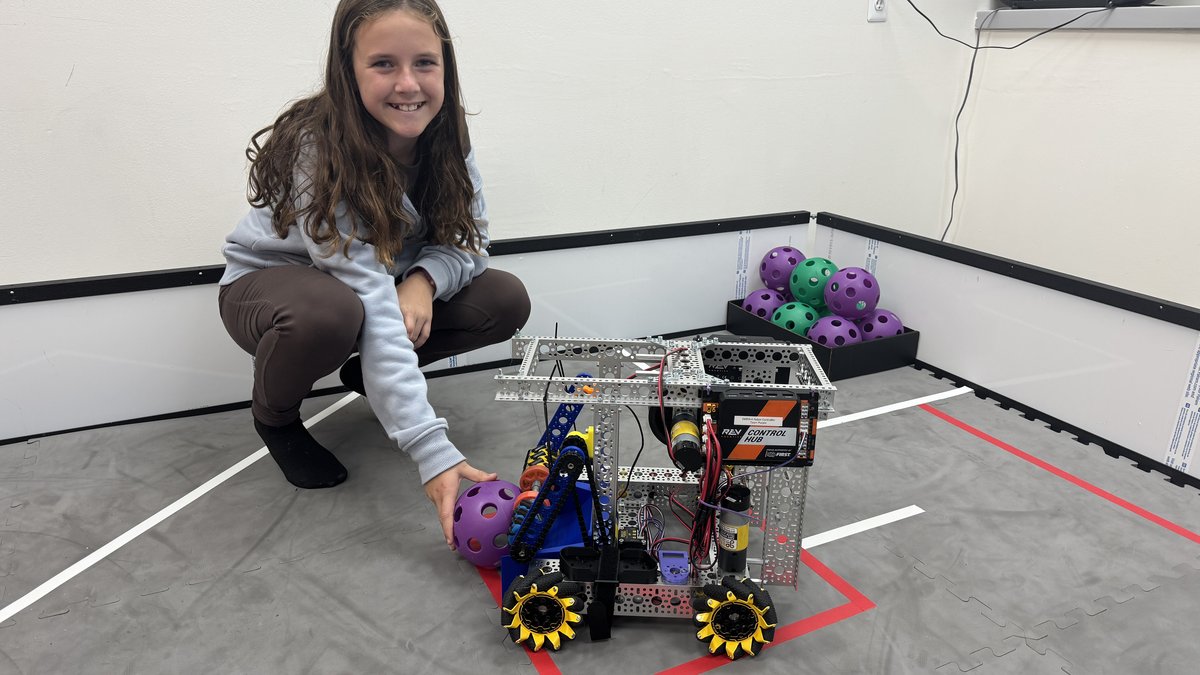 A student smiles as she crouches next to her completed FTC robot on the practice field.